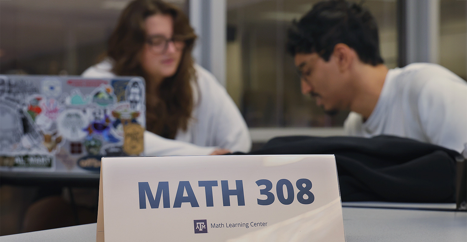 Foreground: Math 308 on a table tent sign. Background: two students studying together